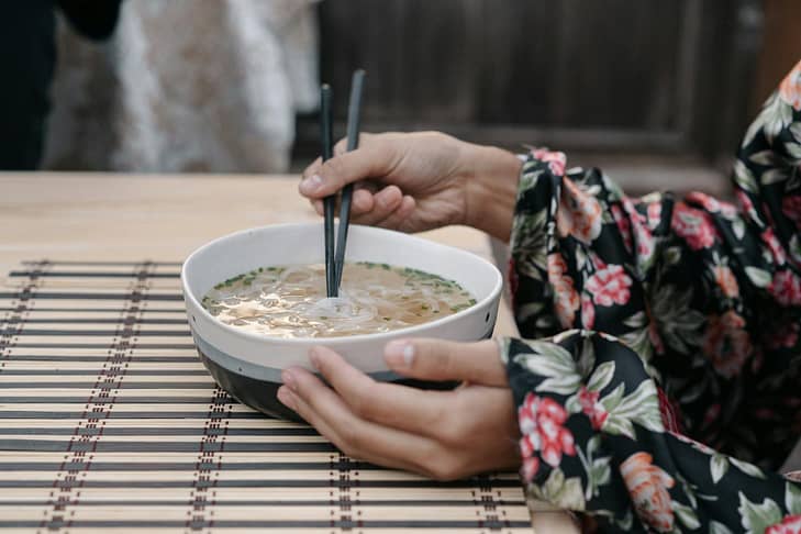 A woman wearing a floral kimono uses chopsticks to eat noodles from a bowl, showcasing traditional dining.